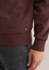 Burgundi spalvos džemperis su rauktomis rankovėmis ir apačia, su mažai juoda logo kairiajame apatinėje dalyje. Audinys atrodo šiek tiek tekstūruotas ir minkštas.