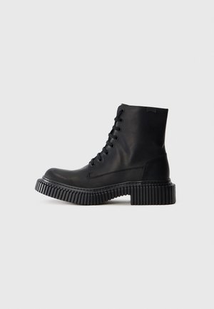 Stivaletto alla caviglia in pelle nera con suola spessa scanalata e chiusura frontale con lacci, mostrato di profilo su uno sfondo grigio chiaro uniforme.