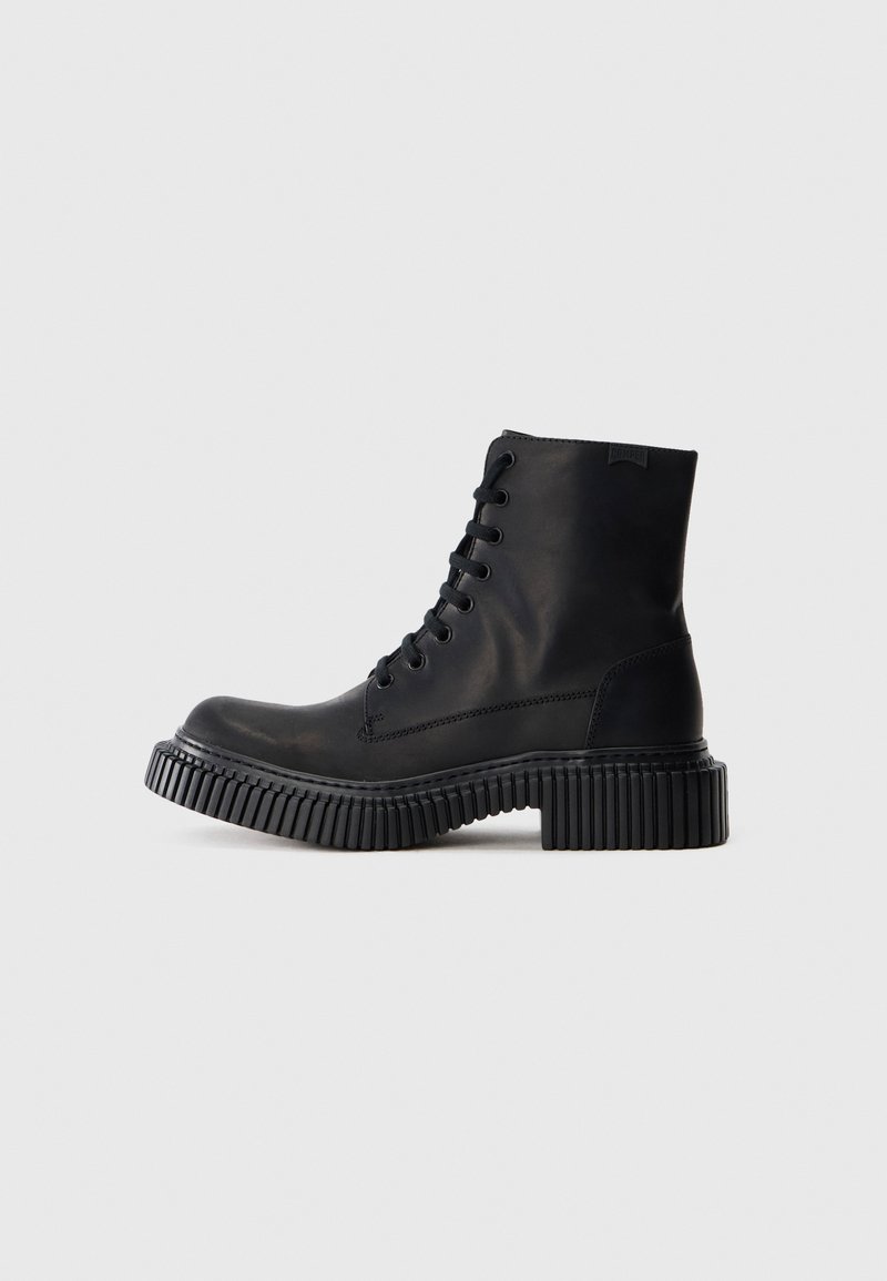 Bottine en cuir noir avec semelle épaisse rainurée et laçage à l’avant, présentée de profil sur un fond gris clair uni.