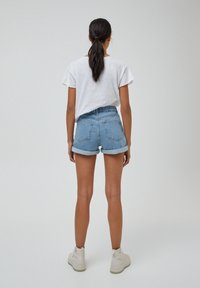 Kvinna med mörkt hår i en hästsvans, klädd i en vit T-shirt, uppvikta ljusblå denimshorts och vita ankelskor, som står mot en enfärgad bakgrund.