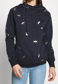 Felpa navy con vestibilità rilassata, decorata con dettagli in oro a forma di libellula e colletto a cowl. Realizzata in tessuto morbido con polsini a coste.