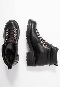 Een paar zwarte leren wandelschoenen met rode en zwarte veters, metalen oogjes en dikke rubberen zolen op een witte achtergrond.