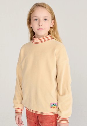 Enfant aux longs cheveux blonds portant un pull beige avec des poignets et un col rayés rouges, et un pantalon rouge, debout sur un fond uni.