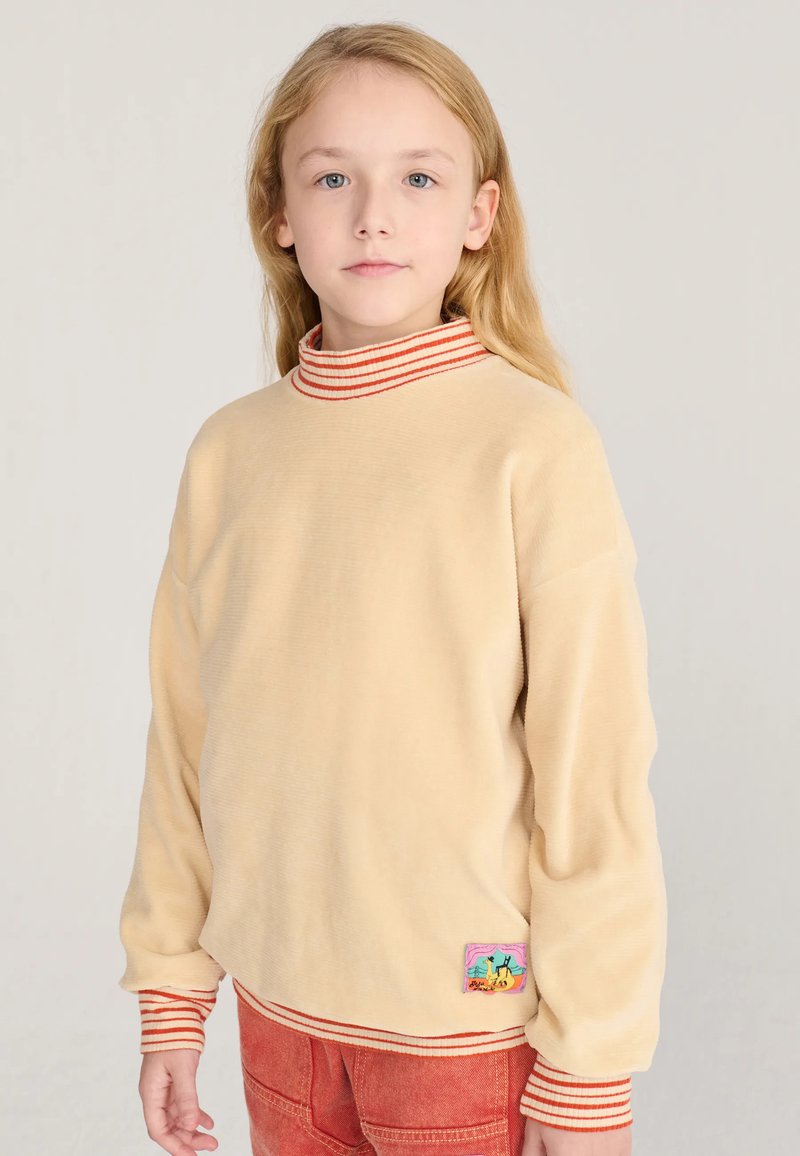 Enfant aux longs cheveux blonds portant un pull beige avec des poignets et un col rayés rouges, et un pantalon rouge, debout sur un fond uni.