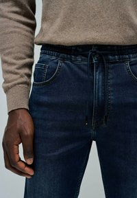 Joggers de mezclilla azul oscuro con cintura elástica y cordón, bolsillos frontales y un ajuste ceñido. El material parece suave y elástico.