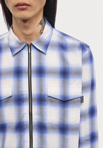 Blaue und weiße karierte Hemd mit Kragen, Reißverschluss vorne, zwei Brusttaschen und einem Stoffetikett. Strukturiertes Muster mit Farbverläufen.