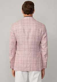 Blazer à carreaux rose clair en tissu texturé, avec une fente unique à l'arrière et des manches ajustées, associé à un pantalon blanc.
