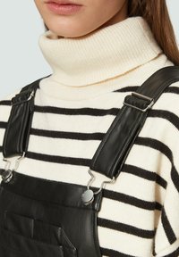 Zwarte leren salopettes met zilverkleurige hardware, gedragen over een crèmekleurige en zwarte gestreepte coltrui, met ribbeltextuur en een voorzak.