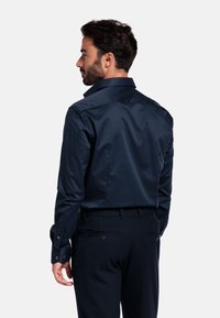 Navyblauw langmouwen shirt met een klassieke kraag. Een gladde textuur en een getailleerd ontwerp. De achterkant toont knoopaccenten op de mouwen en de tailleband.