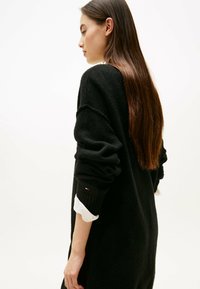 Maglione nero oversize lavorato a maglia con maniche lunghe, con polsini a coste e bordo di una camicia bianca visibile sotto. Texture liscia, design casual.