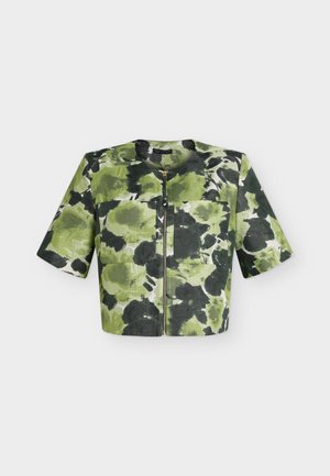 Femme aux cheveux blonds courts porte une veste courte à motif vert et noir par-dessus une chemise blanche, un pantalon blanc et tient un petit sac à main vert.