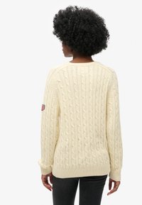 Cremefarbener Strickpullover mit Zopfmuster, geripptem Saum und Bündchen, mit einer Schulterpatch mit Flaggenmotiv. Lockerer Schnitt, strukturierter Stoff, lässiger Stil.