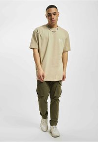 Beige oversize póló rövid ujjal és fehér grafikával, olajzöld cargo nadrággal párosítva, amely több zsebből és rugalmas mandzsettákból áll.