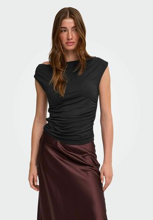 Femme aux longs cheveux bruns portant un haut noir froncé sans manches et une jupe en satin brun brillant, debout devant un fond clair uni.
