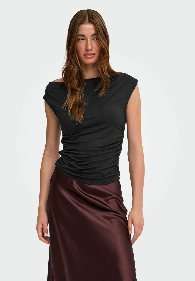 Femme aux longs cheveux bruns portant un haut noir froncé sans manches et une jupe en satin brun brillant, debout devant un fond clair uni.