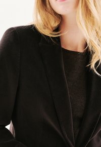 Blazer nero in morbido tessuto con rever a scollatura, sovrapposto a un maglione scuro. Tessuto liscio, forma aderente, senza chiusure visibili.