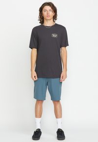 T-shirt en coton noir avec un petit logo sur la poitrine, associé à un short teal. Le modèle porte des baskets noires et des chaussettes blanches à mi-mollet.