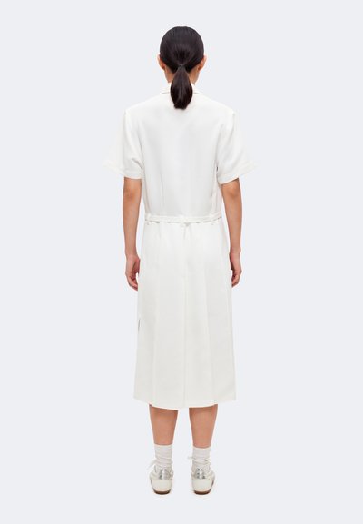 Personne aux cheveux foncés en queue de cheval basse portant une robe midi blanche à manches courtes avec ceinture, chaussettes blanches et chaussures argentées, debout de dos.