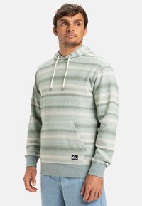 Sudadera con capucha de tela verde suave con rayas blancas horizontales, que cuenta con un bolsillo frontal y capucha ajustable. Logos en el dobladillo.