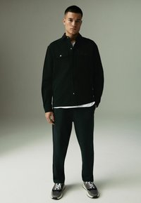 Camisa negra de botones con dos bolsillos en el pecho, llevada sobre una camiseta blanca. Pantalones negros ajustados y zapatillas grises completan el atuendo.