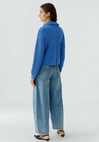 Abgeschnittenes blaues geripptes Sweatshirt mit einem umgeschlagenen Kragen, kombiniert mit locker sitzenden hellblauen Jeans und schwarzen Slippers.