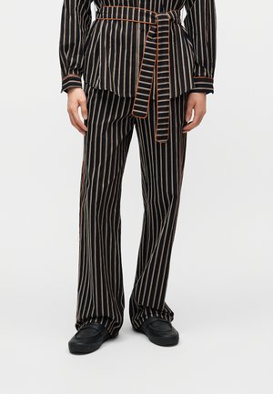 Personne portant un pantalon large noir à jambe droite avec une ceinture assortie à rayures verticales blanches et orange, associé à des chaussures noires sans lacets.