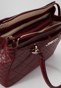 Sac à main matelassé rouge avec une finition brillante, équipé de ferrures dorées, de plusieurs compartiments zippés et d'un extérieur à motif.