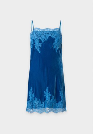 Mujer con cabello largo y oscuro que lleva un vestido de terciopelo azul con ribete de encaje y sandalias negras de tacón, de pie frente a un fondo liso y claro.
