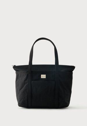 Svart toteveske med doble håndtak, frontlomme, glidelåslukking og liten beige etikett midt foran.