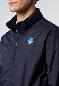 Giacca leggera blu navy con colletto alto e chiusura con zip. Presenta un logo rotondo blu sul petto e una texture liscia e lucida.