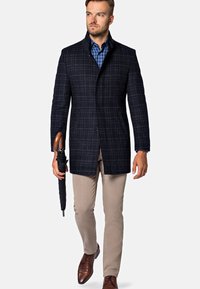 Męski ciemnoniebieski płaszcz w kratę z wyłogami, w połączeniu z koszulą w kratę, beżowymi spodniami, trzymając czarną parasolkę i brązowe buty.