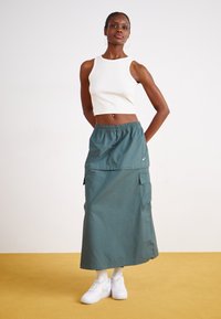 Zöld, bokáig érő szoknya rugalmas derékkal, elülső zsebbel és oldalsó cargo zsebbel. Fehér crop top-pal és fehér sportcipővel párosítva.