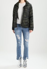 Veste en duvet avec un motif camouflage vert foncé, pull en laine, jeans bleus déchirés avec des ourlets effilochés et bottines métalliques argentées.