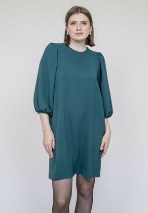 Robe courte couleur sarcelle avec manches bouffantes, encolure ronde et silhouette ample et fluide. Texture lisse avec un léger éclat.