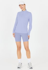 Hellblau-langes Oberteil mit Reißverschluss, kombiniert mit passenden Shorts. Aus weichem, dehnbarem Stoff. Trägt dazu eine weiße Kappe und graue Sportschuhe.