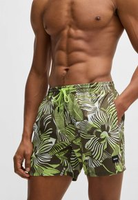 Green and brown patterned swim shorts with a drawstring waistband, featuring palm leaf designs and a small black logo detail.