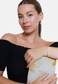 Gouden sieraden met parelaccenten, een delicate armband en een bijpassende ketting, gedragen door iemand die een gestructureerd boek met een gouden design vasthoudt.