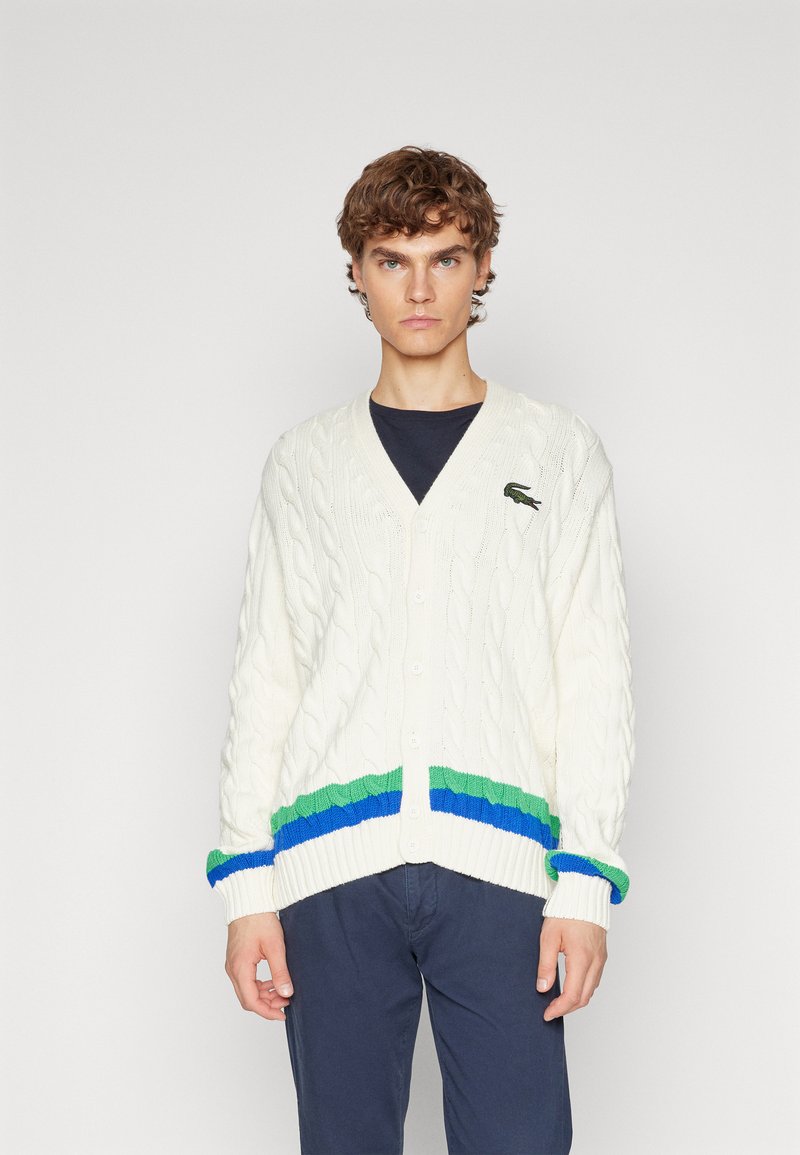 Cardigan en tricot torsadé crème avec des bords côtelés verts et bleus au niveau de l'ourlet et des poignets. Présente un col en V et une fermeture à un bouton.