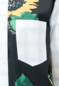 Λευκή τσέπη πουκάμισου με διακριτικό λογότυπο Moschino Jeans σε μαύρο πουκάμισο με πράσινα και κίτρινα αφηρημένα λουλουδένια μοτίβα από κάτω.