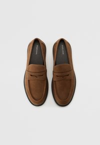 Mocassins en daim marron avec une texture lisse, ornés d'une sangle décorative et de coutures contrastées, avec une semelle en caoutchouc pour une bonne adhérence.