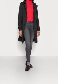 Black trench coat with button detail, worn over a fitted red turtleneck, paired with dark gray jeans and black loafers. Smooth texture throughout.