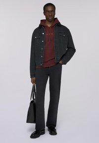 Czarna denimowa kurtka na bordowej bluzie z kapturem ze sznurkami, w połączeniu z ciemnymi spodniami. Trzyma czarną torbę na ramię, włożone błyszczące czarne buty.