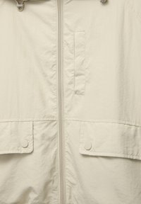 Helle beigefarbene Jacke mit einem Frontreißverschluss, zwei Klapptaschen und einer glatten, leichten Stofftextur. Verstellbarer Kordelzug an der Kapuze.