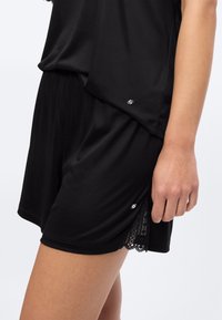 Γυναίκα που φοράει μαύρο φαρδύ σορτς με λεπτομέρεια δαντέλας στο πλάι και ταιριαστό μαύρο τοπ, δείχνοντας τον κορμό και το χέρι της πάνω σε λευκό φόντο.