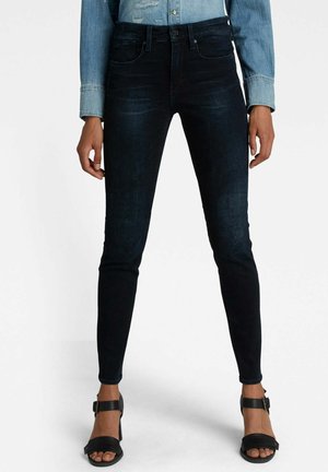 Person wearing dark skinny jeans, black open-toe heels, and a light denim shirt standing with legs slightly apart.