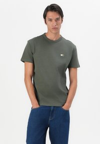 Tricou scurt din bumbac de culoare verde măsliniu, cu guler rotund. Are un mic emblemă imprimată pe pieptul stânga și un croi drept.