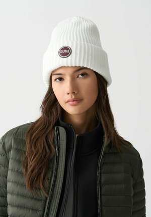 Witte geribbelde gebreide beanie met een zwart rond logo-embleem met de tekst "COLMAR." Het model draagt een donkergroene gewatteerde jas en daaronder een zwart shirt.