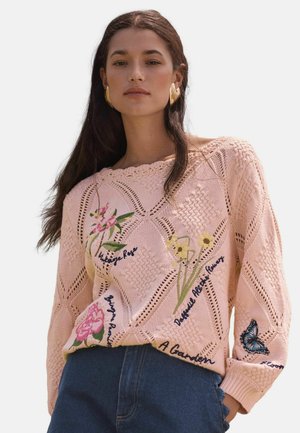Woman with long dark hair wearing a pink knitted sweater embroidered with flowers, butterfly, and text, paired with blue jeans.