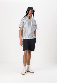 Korte mouwen gestreept shirt in lichtgrijs en marineblauw, gecombineerd met donkere shorts. Gemaakt met een zwarte bucket hat en witte sneakers met tan zolen.