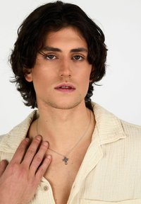 Collana d'argento con pendente a croce nera, esposta su una camicia beige texturizzata. Primo piano del dettaglio del pendente.